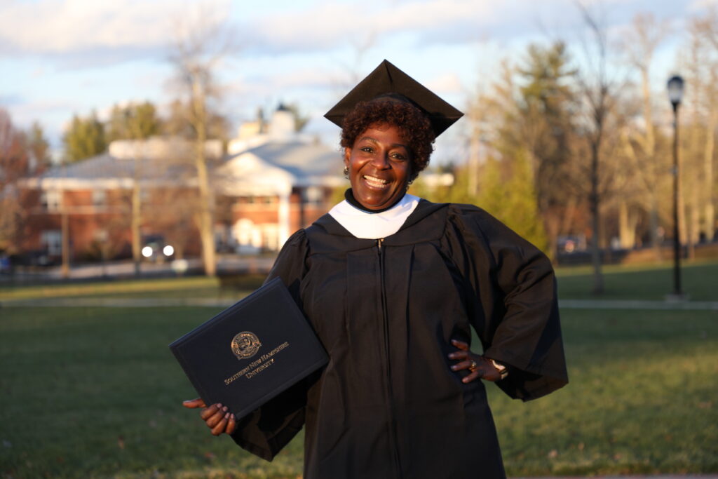 Woman smiling on campus green space, wearing graduation cap and gown, holding diploma cover
